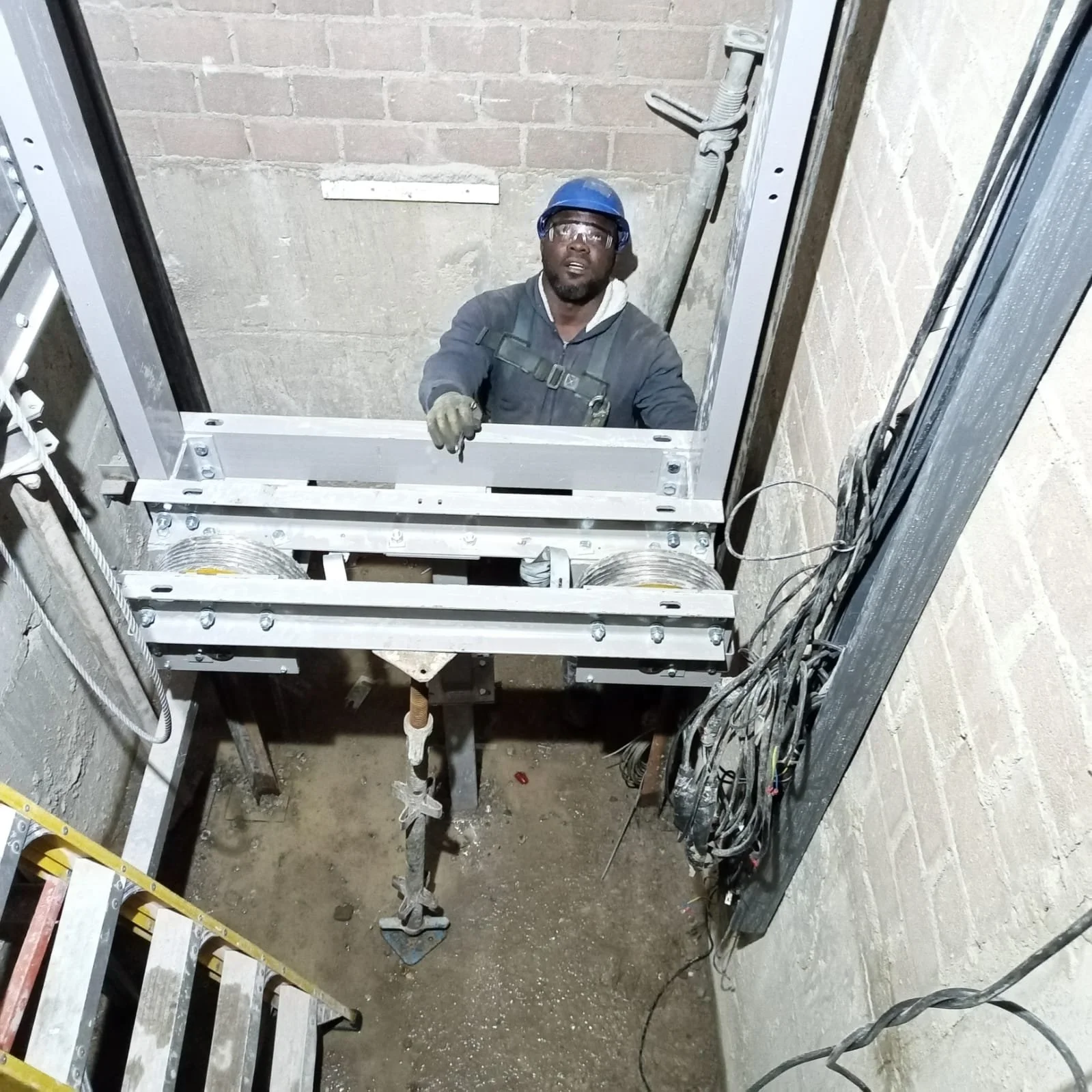 A worker wearing a blue safety helmet, glasses, and gloves is looking up inside an elevator shaft under construction, with metal framework, wires, and concrete walls visible around him.