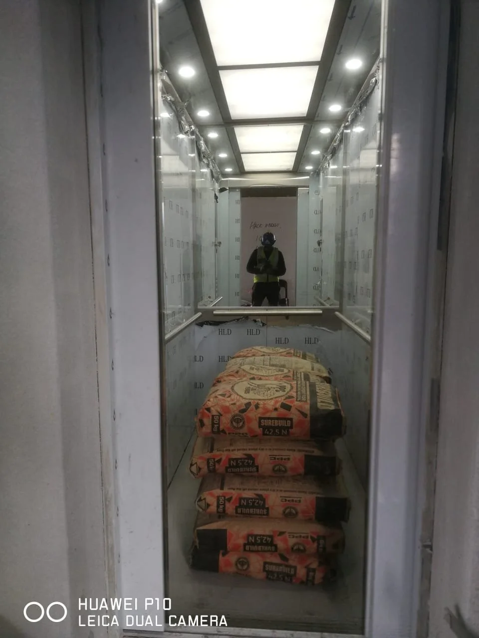 Inside an elevator with construction materials on the floor, a worker in a safety vest and helmet taking a photo of their reflection in the elevator mirror.