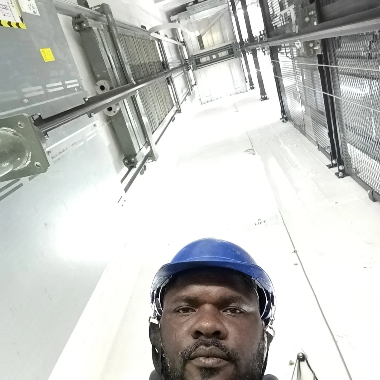 Self-portrait of a man wearing a blue helmet, taking a selfie from a low angle, in an industrial or construction setting with metal structures and a lift.