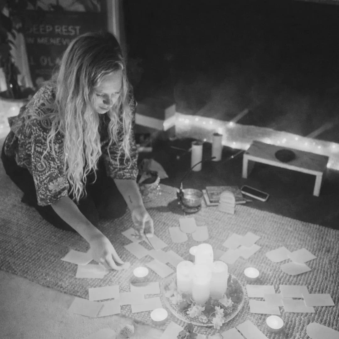 A woman with long, wavy hair sitting on the floor, arranging candles, cards, and flowers in a circle around a cluster of candles on a tray. The room is dimly lit with some fairy lights and a sign that says "Keep Rest" in the background.