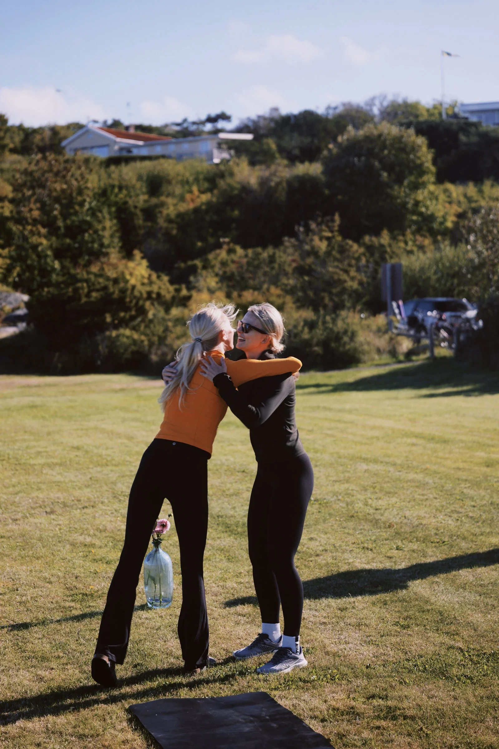 Two women hug and smile outdoors on a grassy area, with trees, a parking lot, and a hillside with houses in the background.