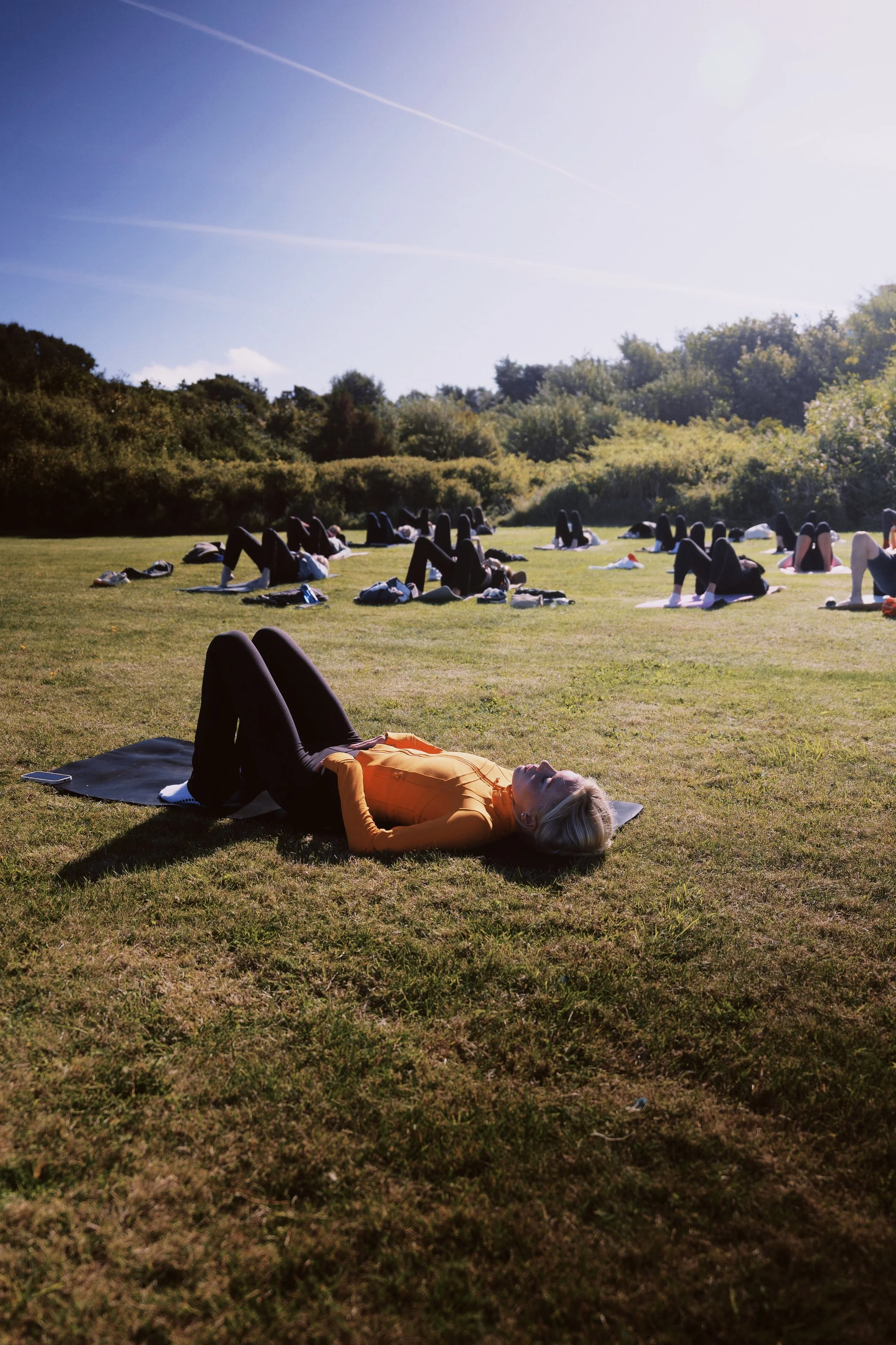 A woman lying on a yoga mat outdoors in a grassy field with other people practicing pilates in the background.