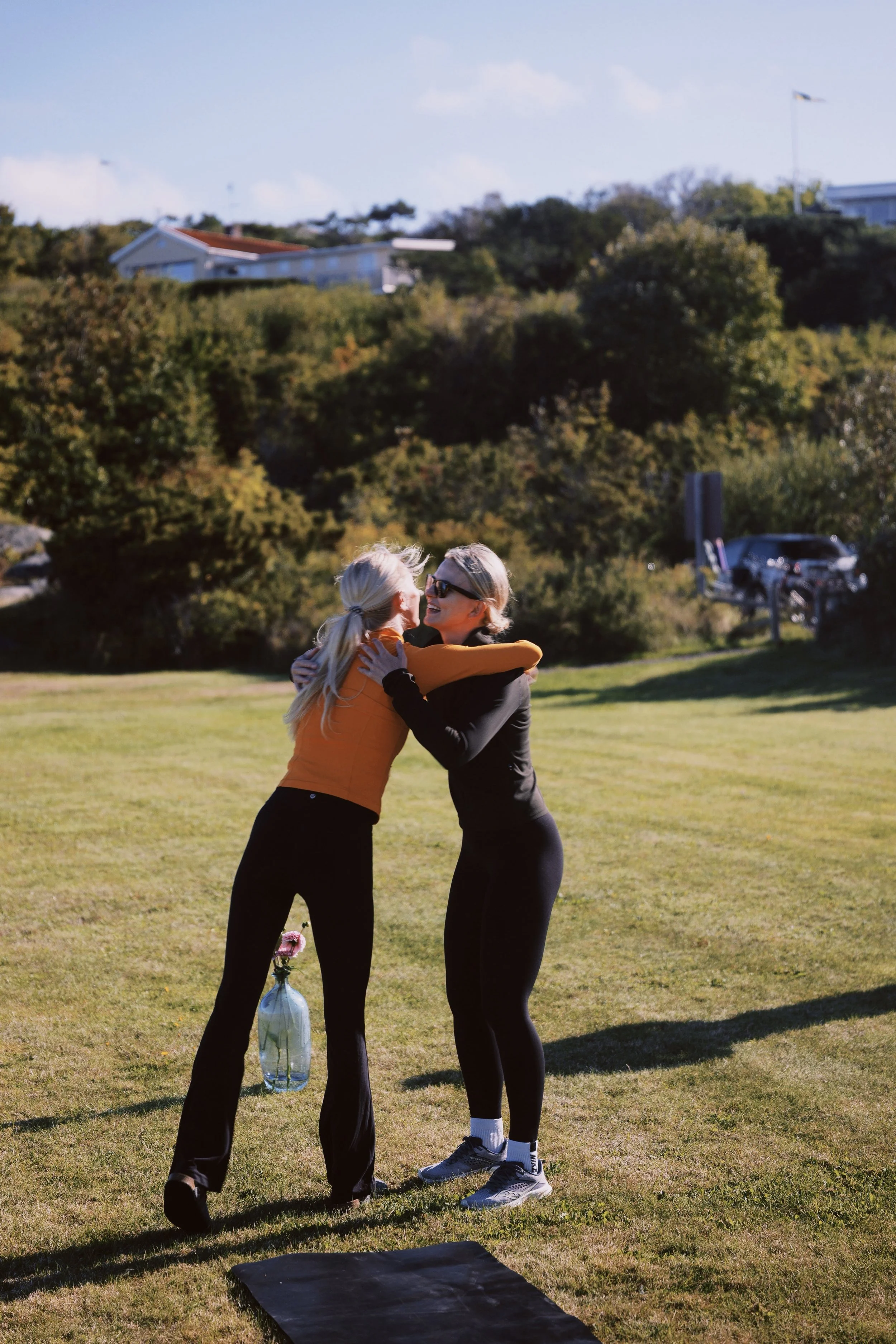 Two women embracing and laughing outdoors on a grassy area, with trees, buildings, and parked cars in the background.