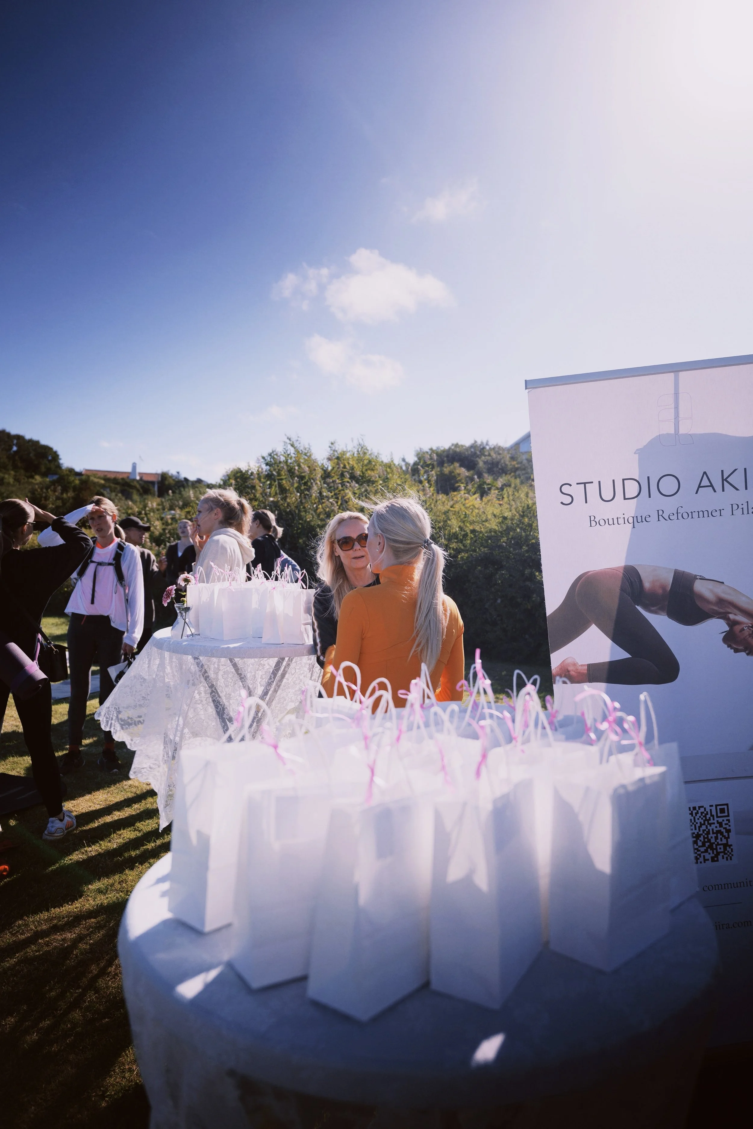 Outdoor event with a group of people gathered around a table with white gift bags and pink ribbons, bright sunny sky with clouds, and a sign reading "STUDIO AKI Boutique Reformer Pilates".