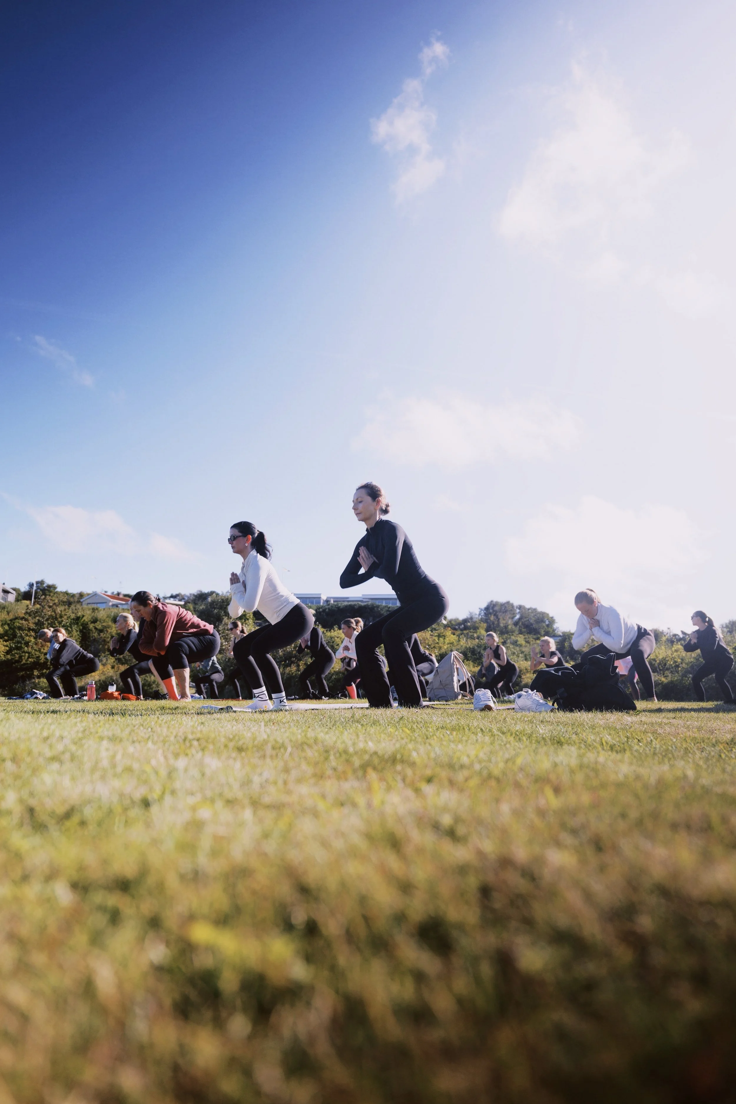 Multiple people practicing pilates outdoors on a grassy field on a sunny day, with blue sky and scattered clouds in the background.