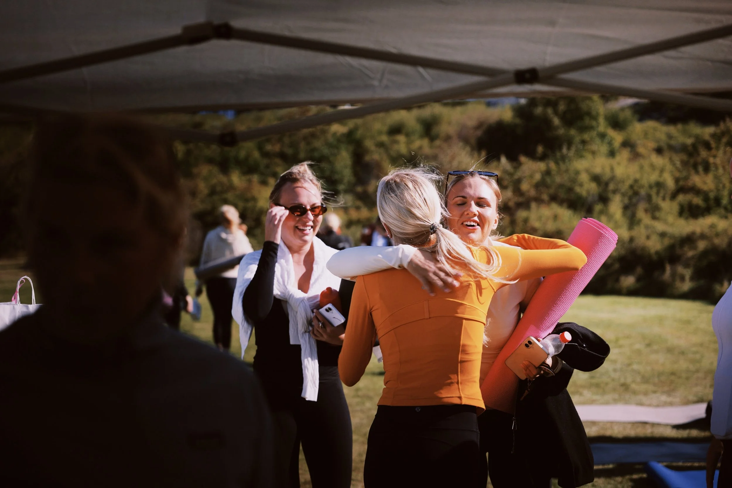 People gathered outdoors, with some wearing sunglasses; woman in orange holding a yoga mat, other individuals smiling and chatting.