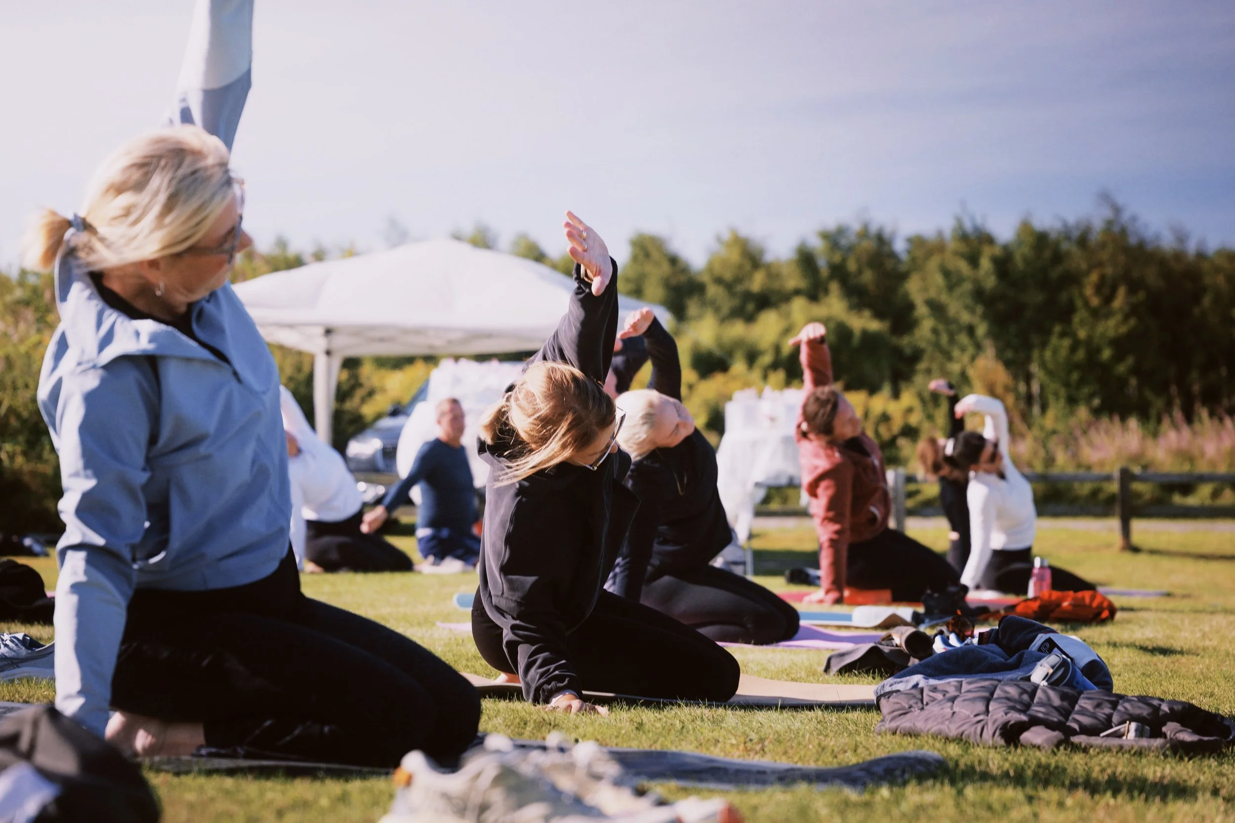 Group of people participating in outdoor pilates session on grass, with some raising their arms and others in kneeling positions, outdoors on a clear day.