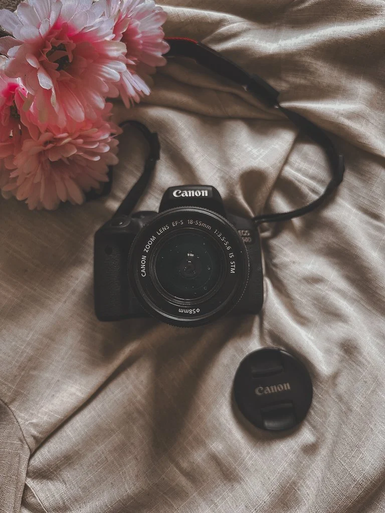 A Canon digital camera with an 18-55mm lens, a lens cap nearby, and pink flowers on a beige fabric surface.