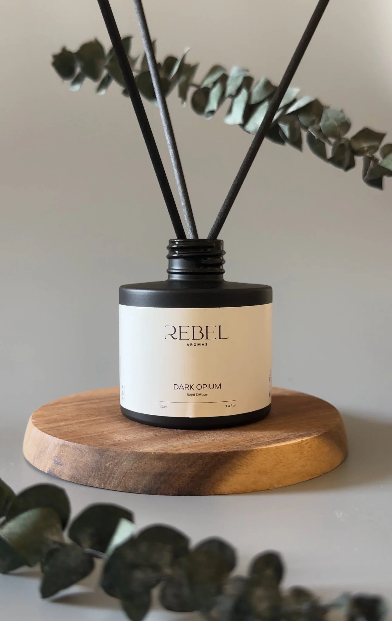 A black and white labeled reed diffuser on a wooden surface, with green eucalyptus leaves in the background.