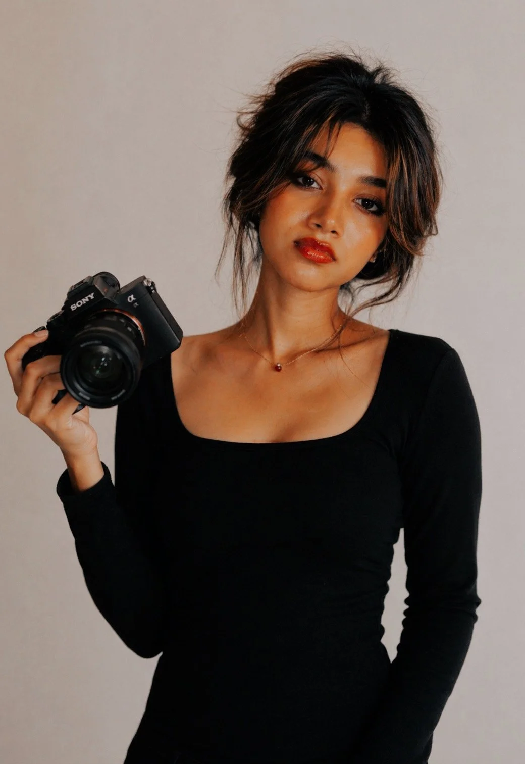 A woman with dark, wavy hair and red lipstick, wearing a black top and a necklace, holds a Sony camera in her right hand against a plain wall background.