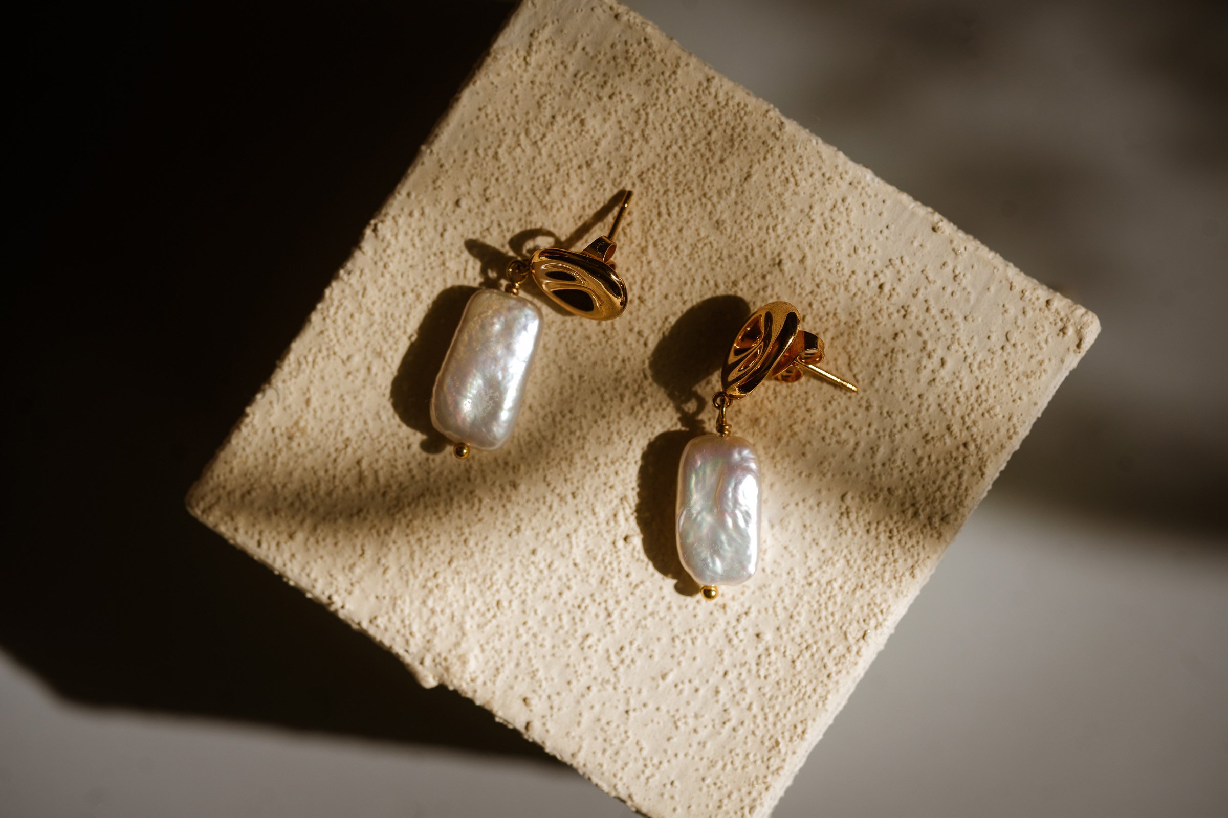 Gold earrings with large irregular pearl pendants displayed on a textured beige foam block.