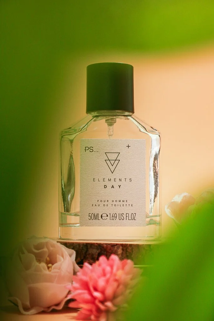 A clear glass perfume bottle with a black cap, labeled "Elements Day Eau De Toilette," sitting on a wooden surface surrounded by pink and white flowers with green leaves in the foreground and background.
