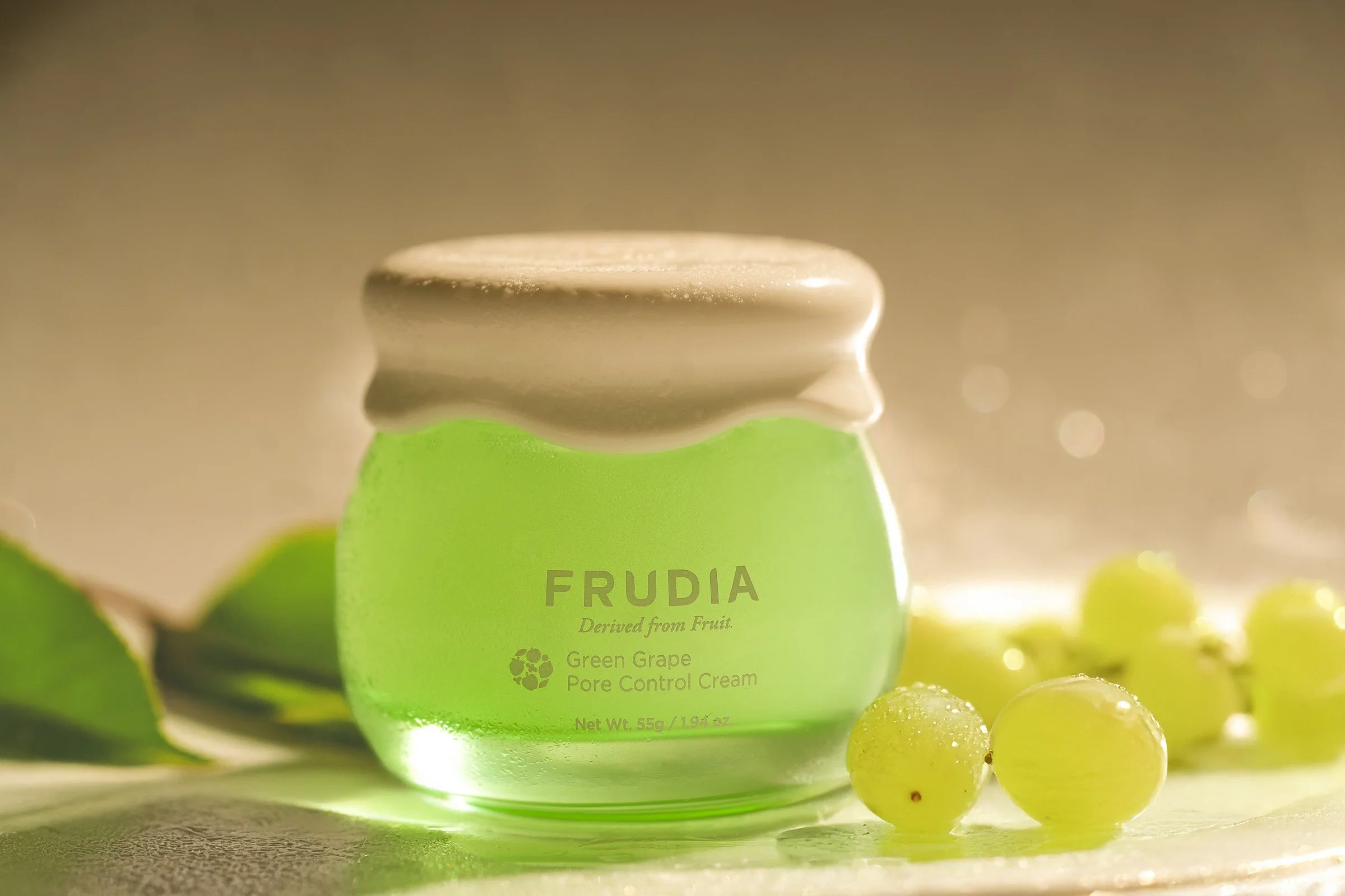 Close-up of a small green jar labeled 'FRUDIA' with cream on the lid, surrounded by green grapes.