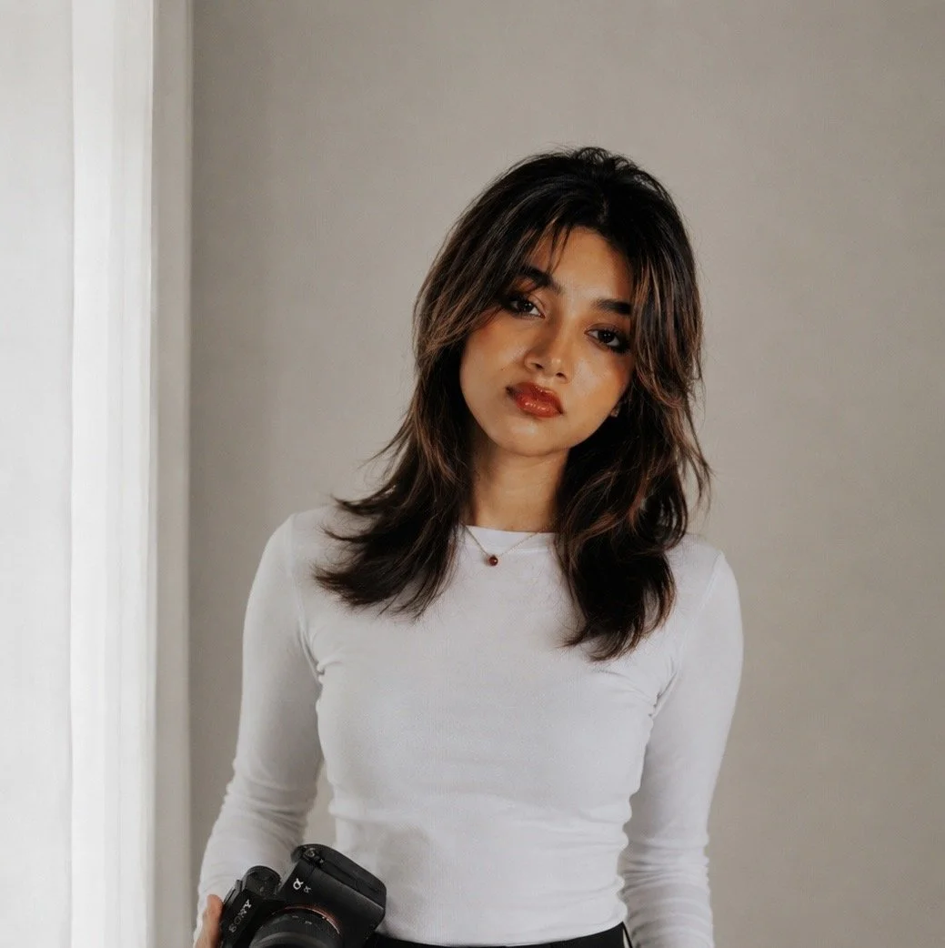 A young woman with wavy brown hair holding a camera, wearing a white long-sleeve shirt, standing in front of a plain gray wall.