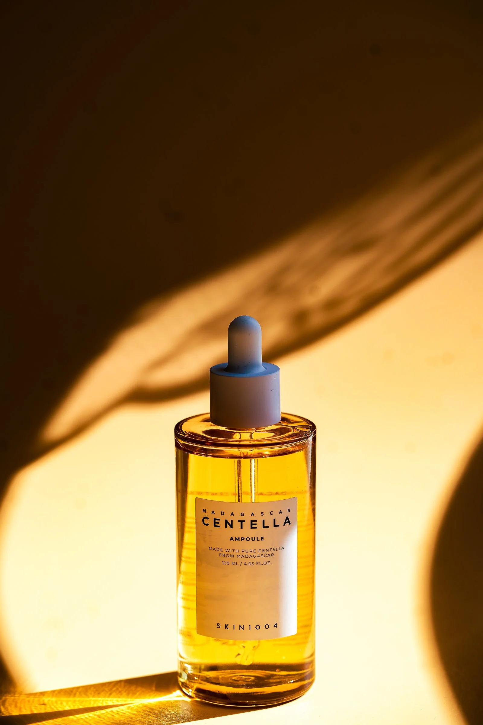 A clear bottle of Madagascar Centella ampoule with a white dropper cap, set against a warm yellow light, casting a shadow on the background.