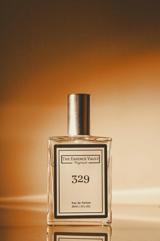 A clear glass perfume bottle with a silver cap, labeled 'The Essence Vault Fragrances 329, Eau de Parfum 30ml.' The bottle is placed on a reflective surface with warm lighting.