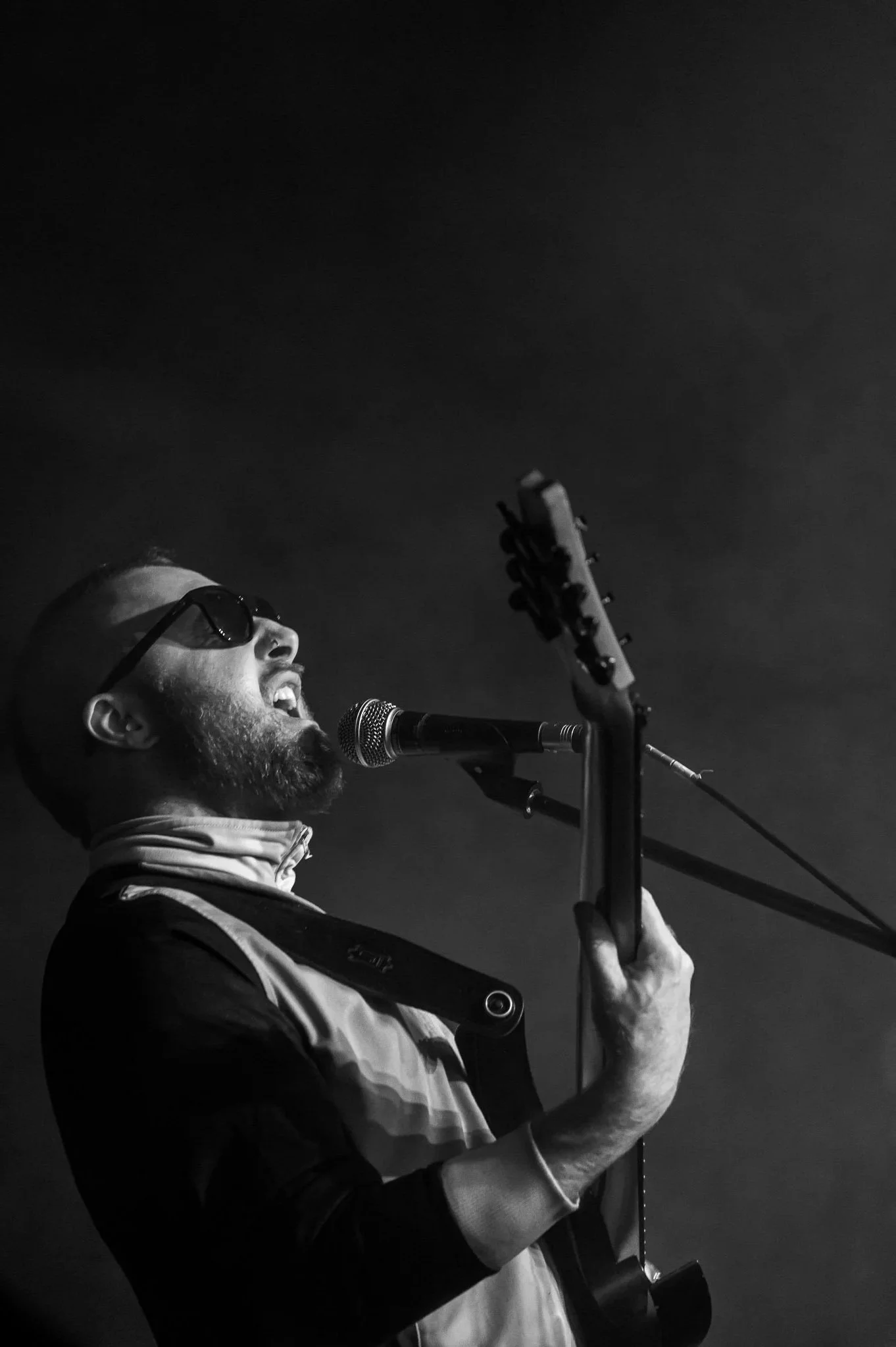 Della Nova, 15th, May, 2026. A black and white photo of a man wearing sunglasses, singing into a microphone, and playing an electric guitar on stage.