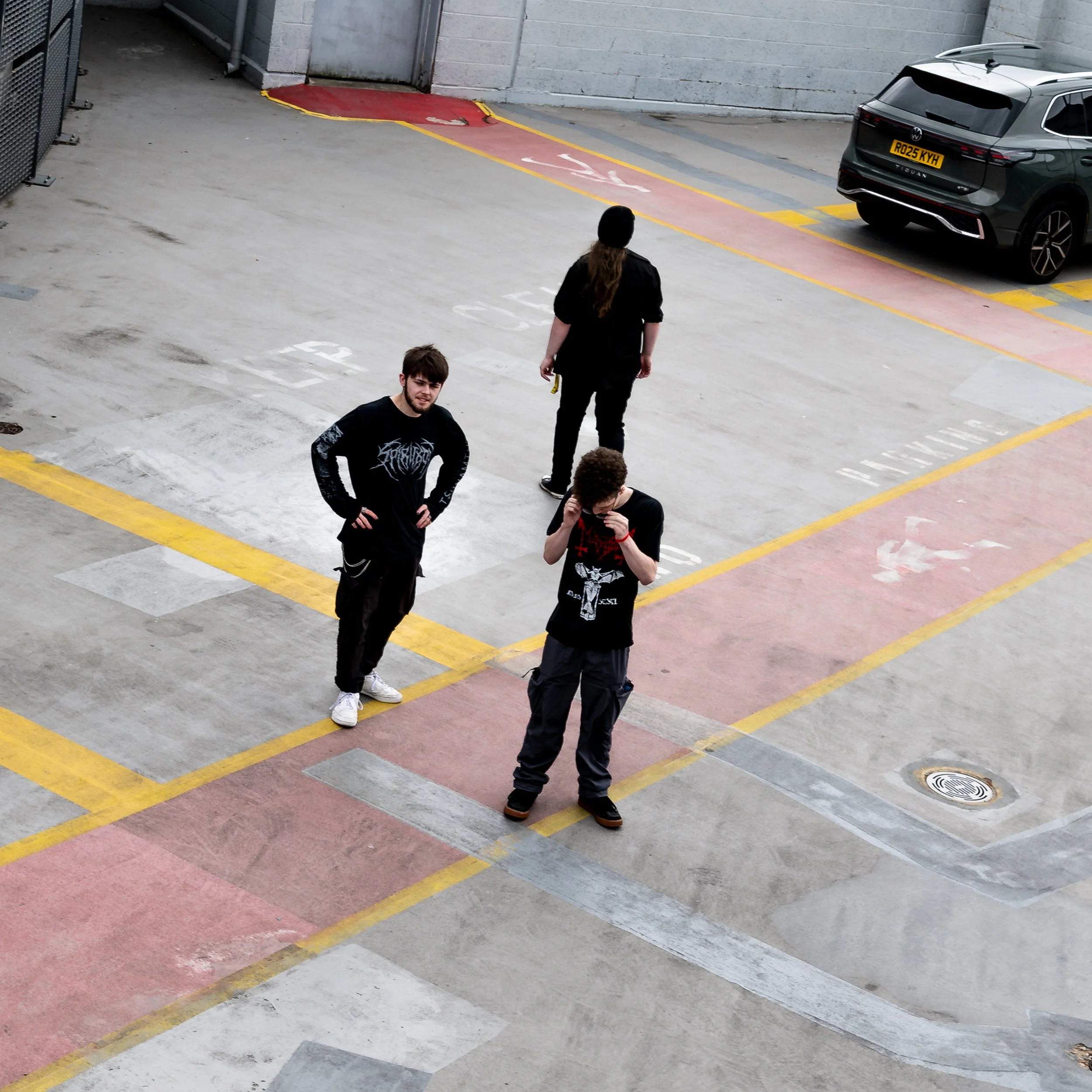 A parking lot with yellow lines and a red painted lane that has white arrows indicating direction. Four people are standing in the parking lot, with one person in the foreground wearing a black shirt with a graphic design. A woman is walking away from the camera near a gray SUV with a yellow license plate. The concrete walls and a closed metal door are visible in the background.