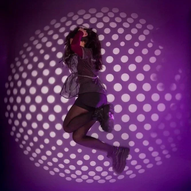 Studio Shoot, 16th, April, 2026. A woman lying on a circular pattern of purple and white lights, wearing a dark outfit with stockings and boots, in a dance or playful pose.