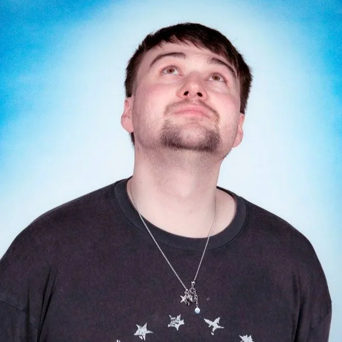 A man with light skin, brown hair, and a goatee looking upward against a gradient blue to white background, wearing a black t-shirt with white star and bird designs and a silver necklace.