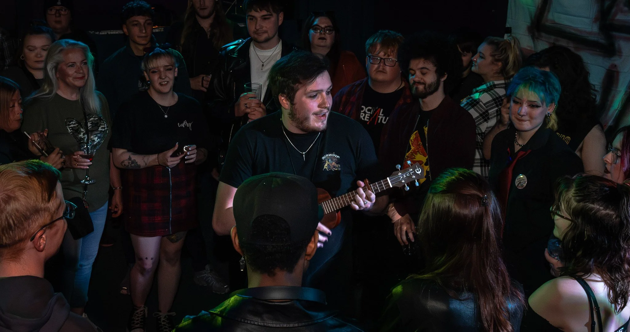 Phoenix Last Stand, 23rd, May, 2025. A crowd of people gathered around a man playing a ukulele in a dimly lit setting, possibly a bar or club, with colorful lighting effects.