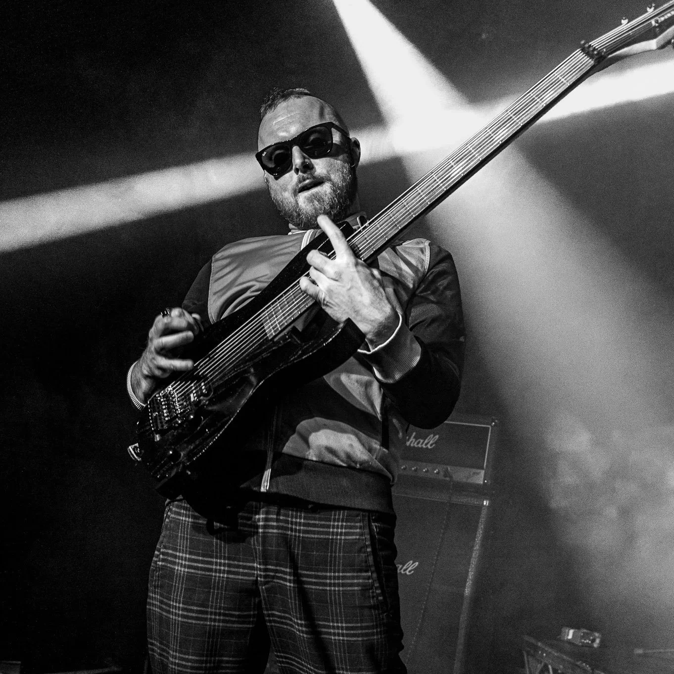 Della Nova, 9th, May, 2026. A man playing an electric guitar on stage, wearing sunglasses, a plaid skirt, and a jacket with a color blocking design, with stage lights in the background.
