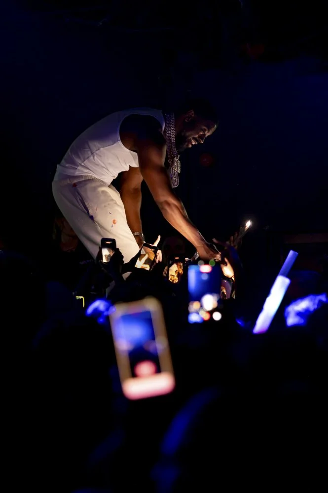 Tion Wayne, Bucks Weekender, 3rd May, 2026. A performer on stage at a concert, dressed in white, wearing jewelry, and interacting with the audience using social media on his phone amidst crowd phones recording.
