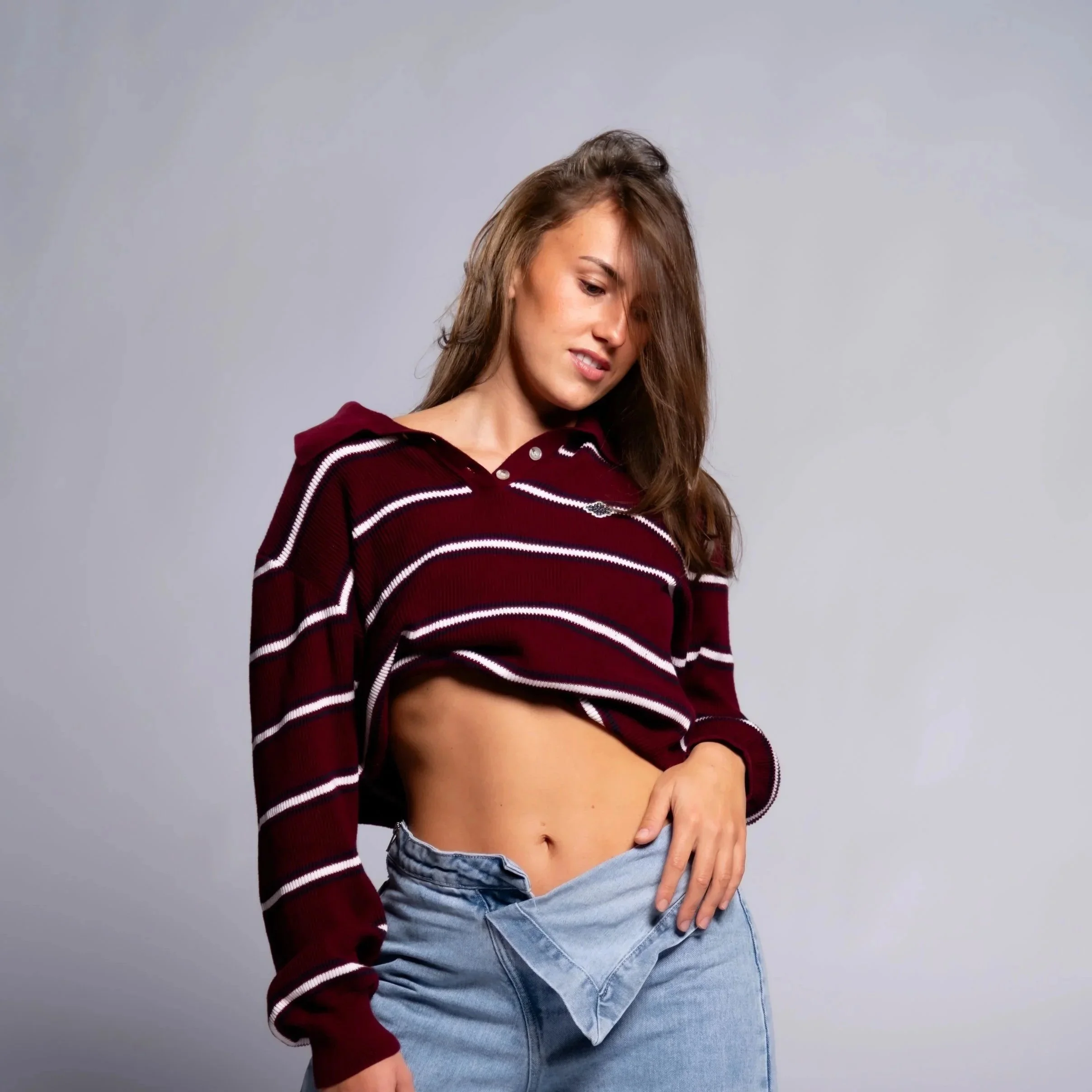 JayLee Jones, Studio Shoot, 14th November, 2026. A young woman with brown hair wearing a maroon and white striped hoodie and light blue jeans, standing against a gray background, with one hand resting on her hip.