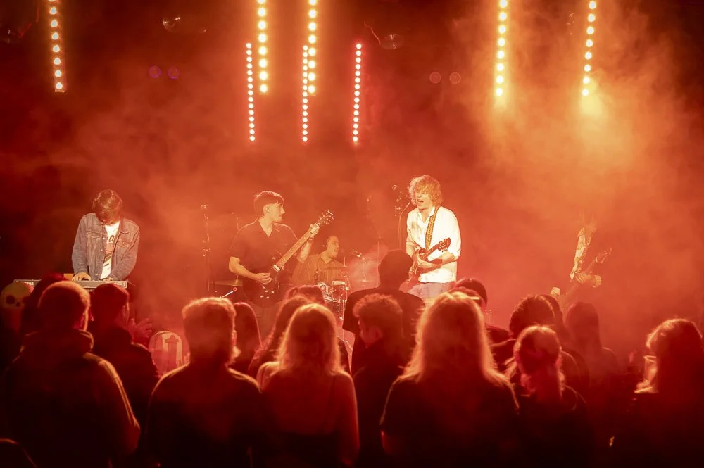 Katemba, Band Slam New Legacy, 12th May, 2026. A band performing live on stage with orange stage lights, featuring a keyboard player, a guitarist, a drummer, and a vocalist. Audience seated watching the show.