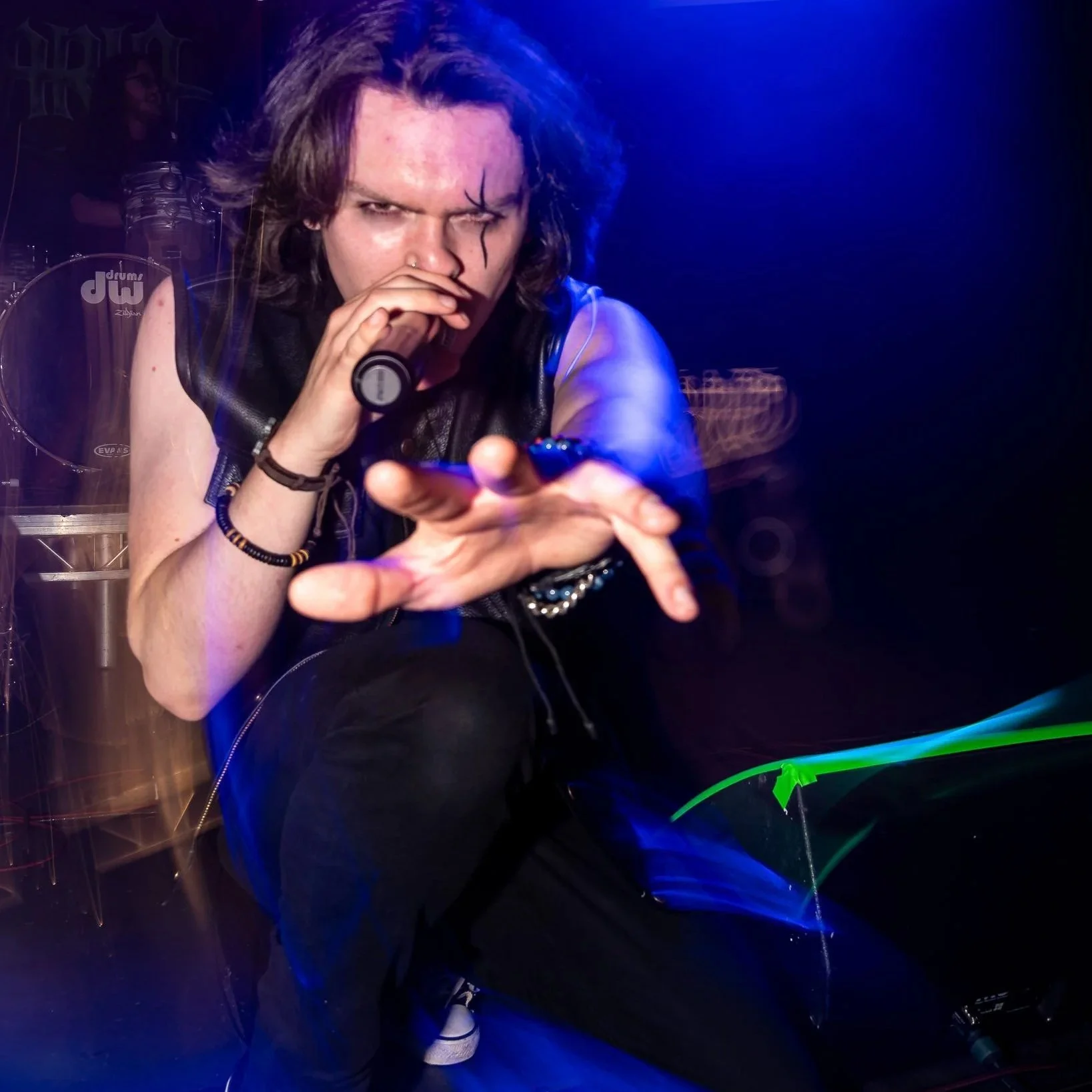 Calvariaband, 15th, May, 2025. Person with long hair, wearing a sleeveless black leather vest, holding a microphone, performing on stage with drums in the background, and dramatic blue and purple stage lighting.