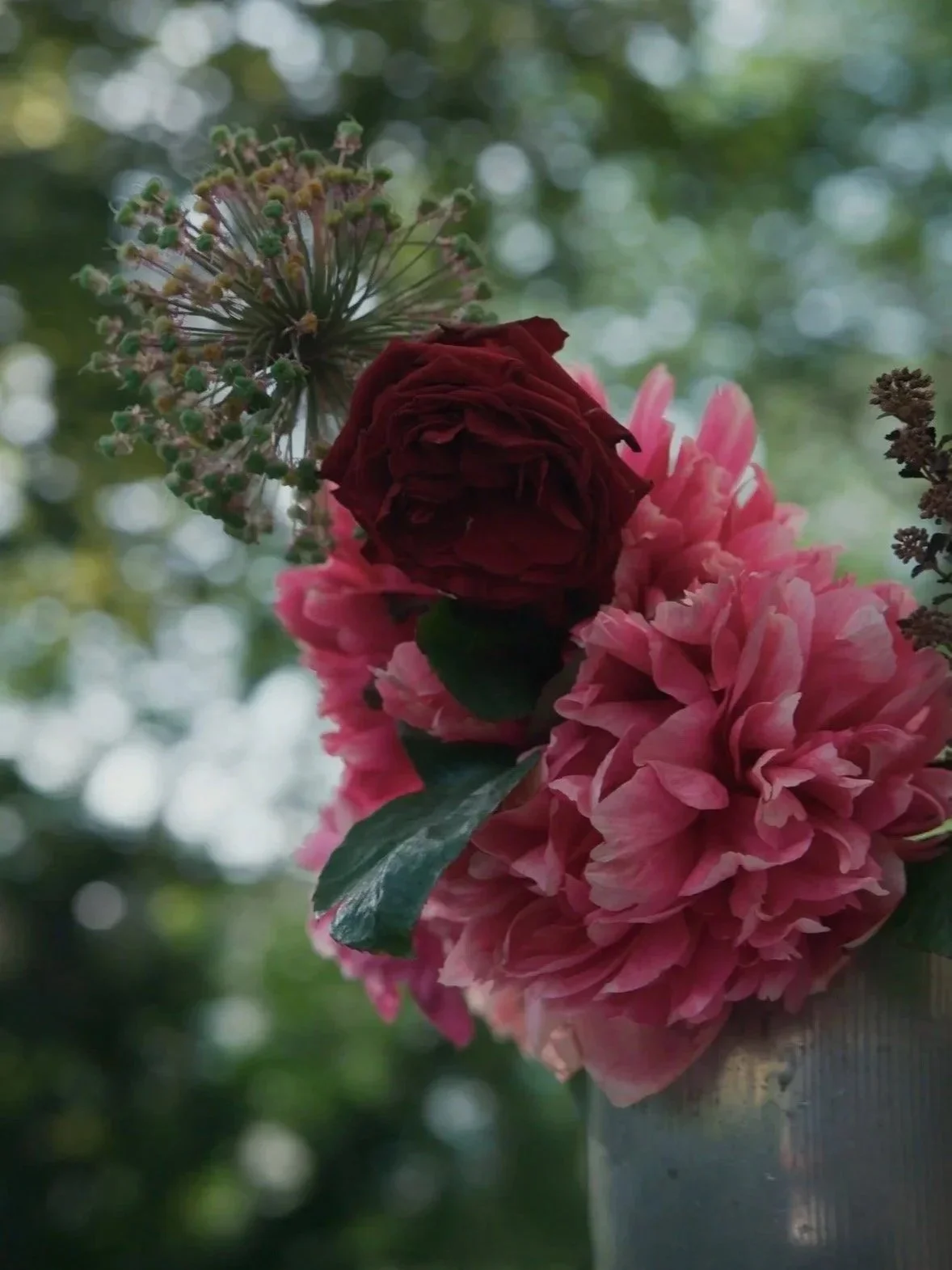 Blumenstrauß mit roten Rosen, pinken Pfingstrosen und einem Hanglampenähnlichen grünen Blumenkeim, vor einem unscharfen Hintergrund aus grünen Blättern.