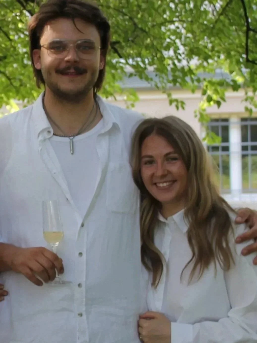 Ein Mann und eine Frau lachen und posieren im Freien, sie tragen weiße Hemden, im Hintergrund sind grüne Bäume und eine moderne Gebäude-Fassade sichtbar.