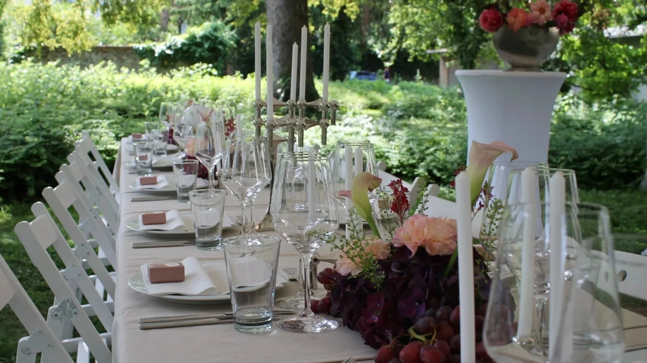 Ein festlich gedeckter Gartentisch mit weißem Tischtuch, Blumenarrangements, Kerzen, Gläsern und Besteck, im Grünen bei Tageslicht.