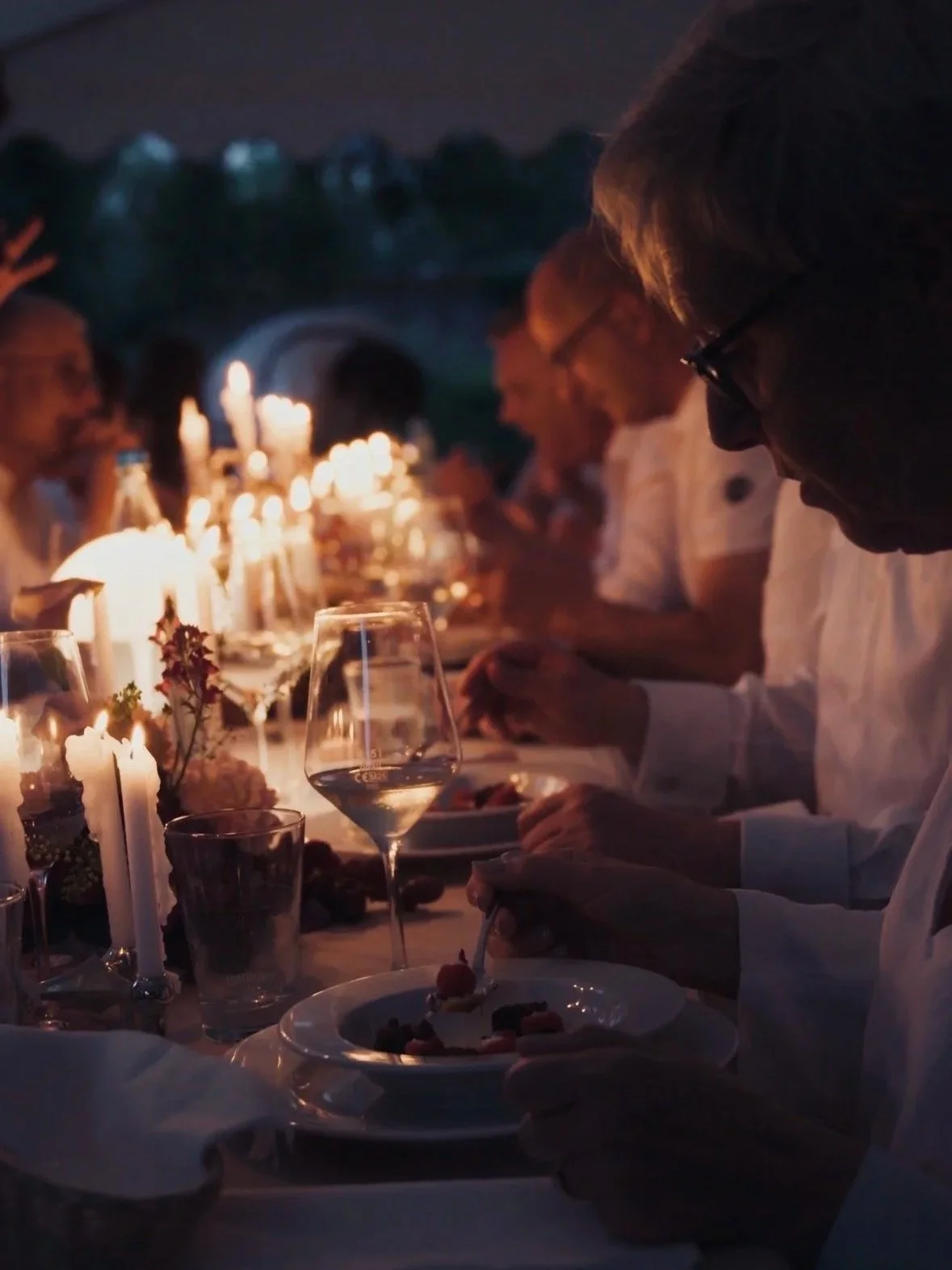 Menschen sitzen bei Nacht an einem festlich gedeckten Tisch mit Kerzen, essen und trinken in einer eleganten Atmosphäre.