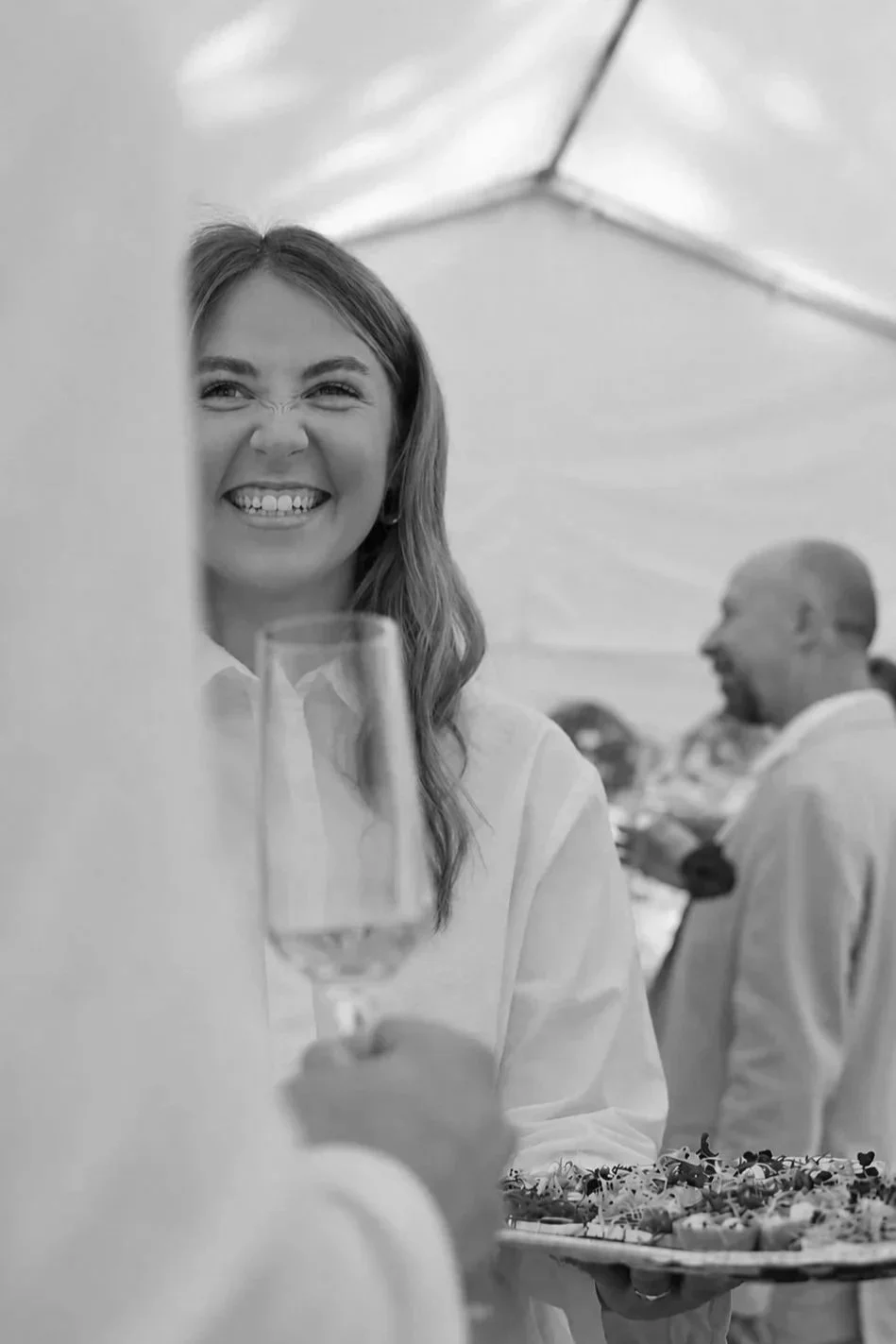 Eine lachende Frau mit langem Haar hält ein Glas Sekt und einen Teller mit Essen bei einer Feier im Freien, im Hintergrund sind weitere Gäste unter einem Zelt.