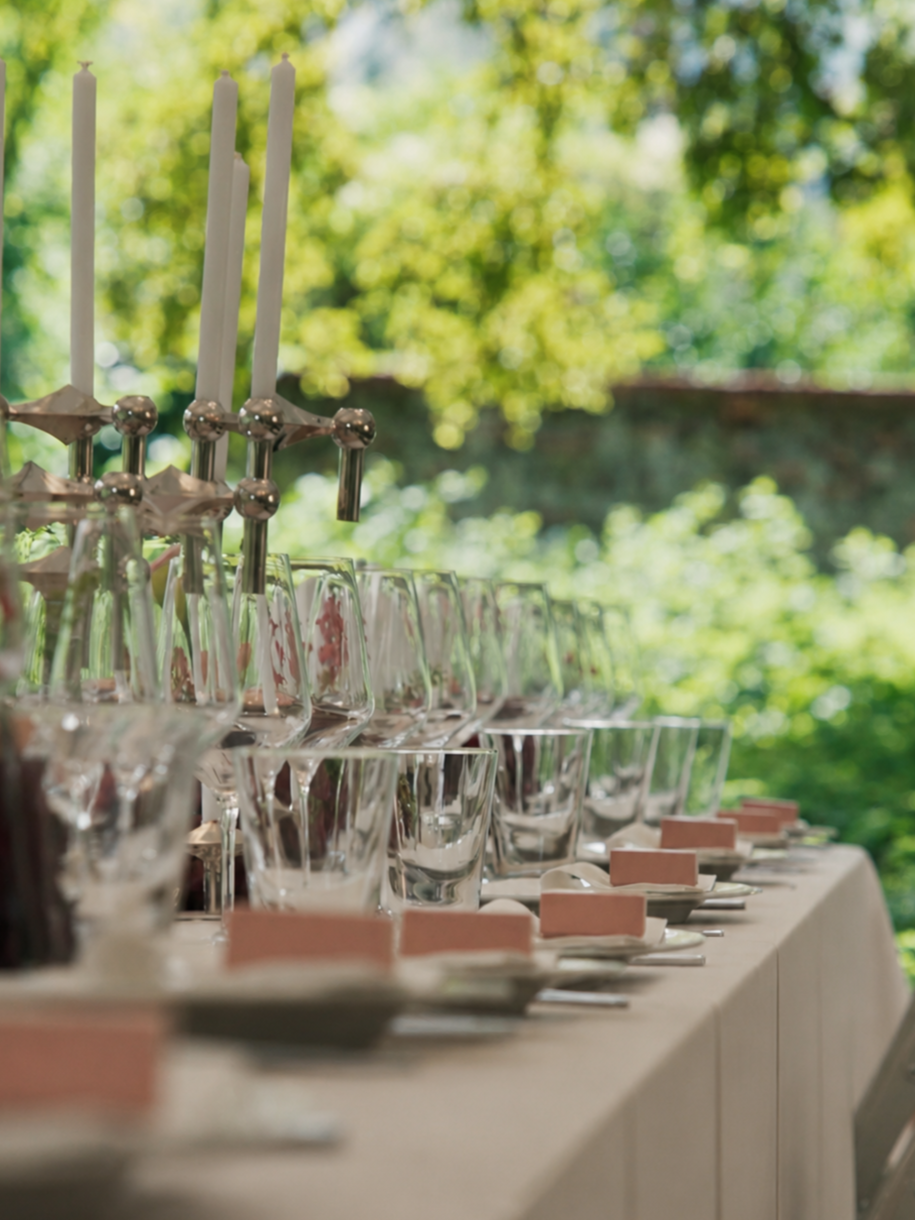 Ein festlich gedeckter Tisch mit leeren Weingläsern, weißen Tellern und rosa Platzkarten, im Hintergrund eine Kerzenhalter mit weißen Kerzen, vor grüner Natur