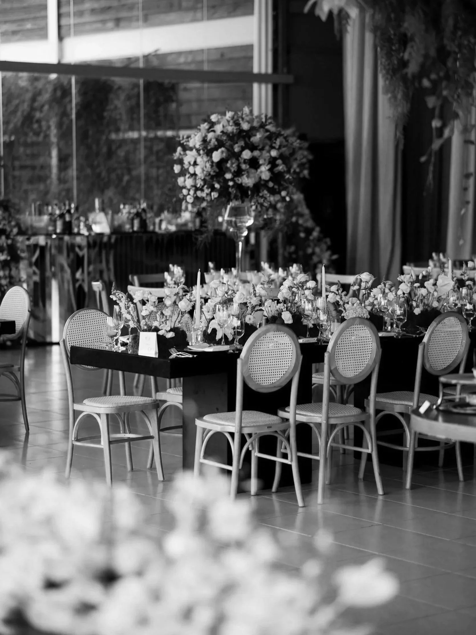Ein eleganter Veranstaltungsraum mit Tischen, die mit Blumenarrangements, Kerzen und Gläsern dekoriert sind. Im Hintergrund befindet sich ein großer Blumenstrauß auf einem hohen Ständer.