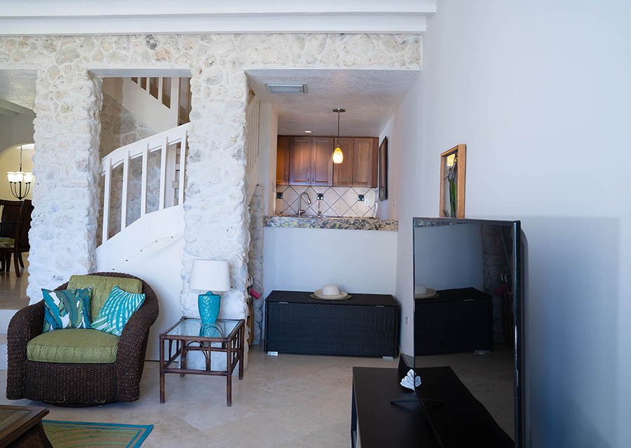 Living room area with a brown wicker armchair with green and blue pillows, a small wooden side table with a white lamp and blue vase, a flat-screen TV on a stand, and a view into a kitchen with wooden cabinets. Stone accents and white walls. There is