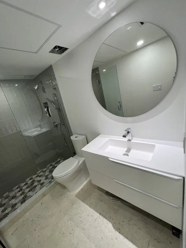 Modern bathroom with a glass-enclosed shower, a white toilet, a white vanity with a rectangular sink, a large round mirror, and minimalistic design.