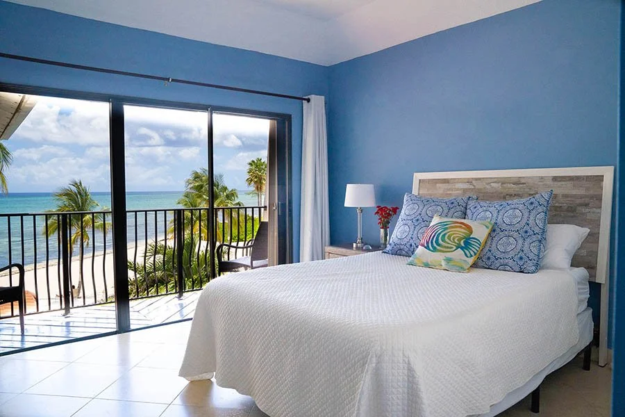 Bedroom with blue walls, white bedding, decorative pillows, a nightstand with a lamp and flowers, and a sliding glass door leading to a balcony with tropical ocean view and palm trees.