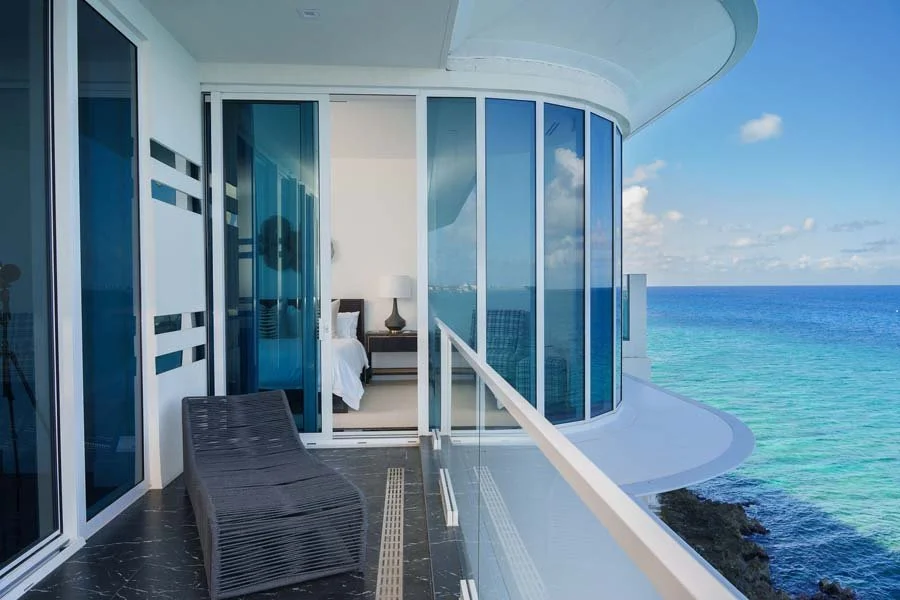 Balcony of a modern high-rise apartment overlooking the ocean, with a black lounge chair and sliding glass door leading to a bedroom.