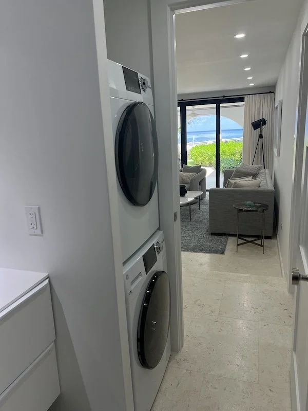 Part of a laundry area with stacked washer and dryer, view into a living room with sofa, armchair, side table, painting, floor lamp, and a large sliding glass door with outdoor view.