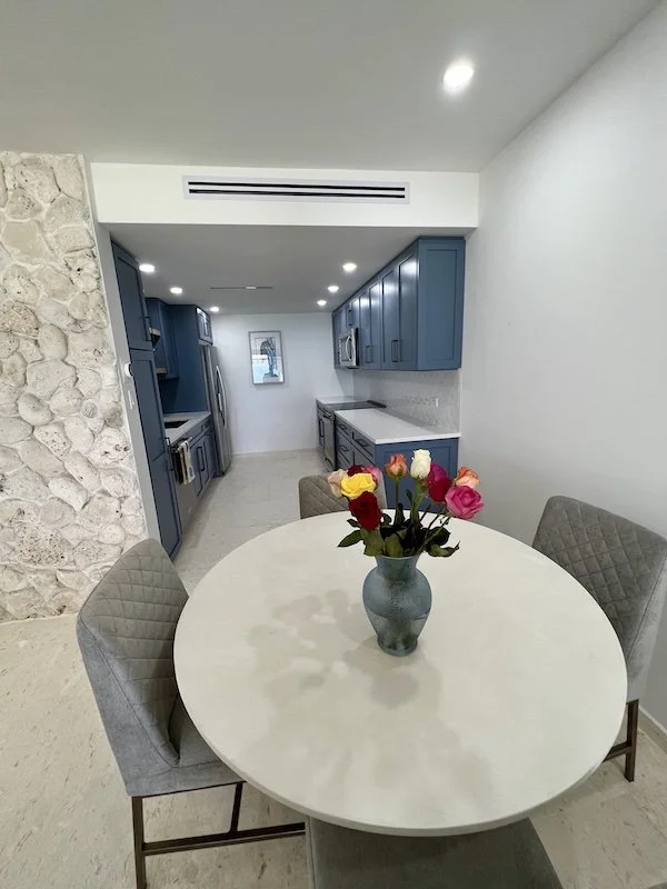 A dining area with a white oval table, a gray vase with colorful roses, and three gray upholstered chairs, with a blue kitchen and stone accent wall in the background.