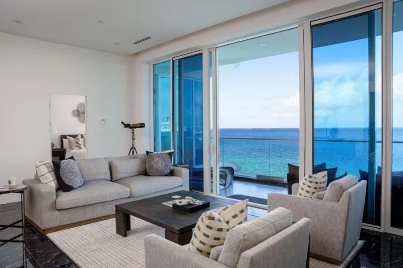 Living room with large sliding glass doors and ocean view, beige sofa, armchairs, black coffee table, telescope, and modern decor.
