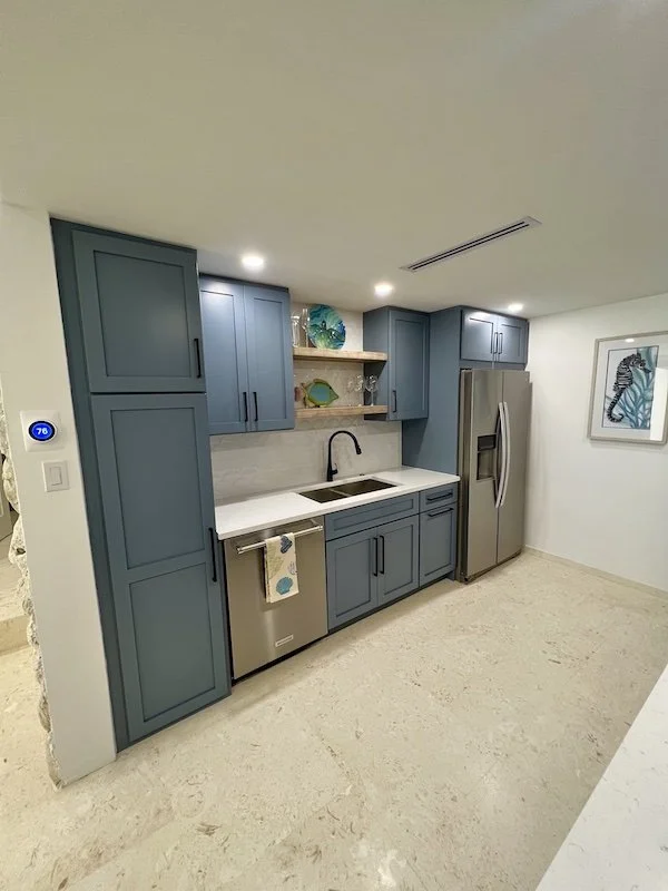 Kitchen with blue cabinets, a stainless steel refrigerator, a white countertop with a sink, open shelves with decorative glassware, and wall art featuring a seahorse.