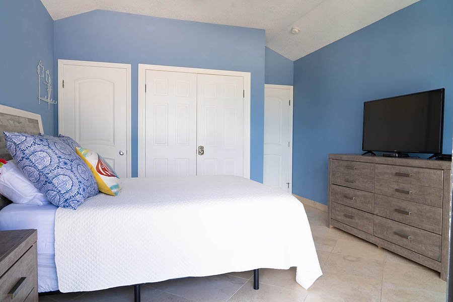 A bedroom with blue walls, a white bed with colorful pillows, a wooden dresser with a TV on top, and two closed white closet doors.