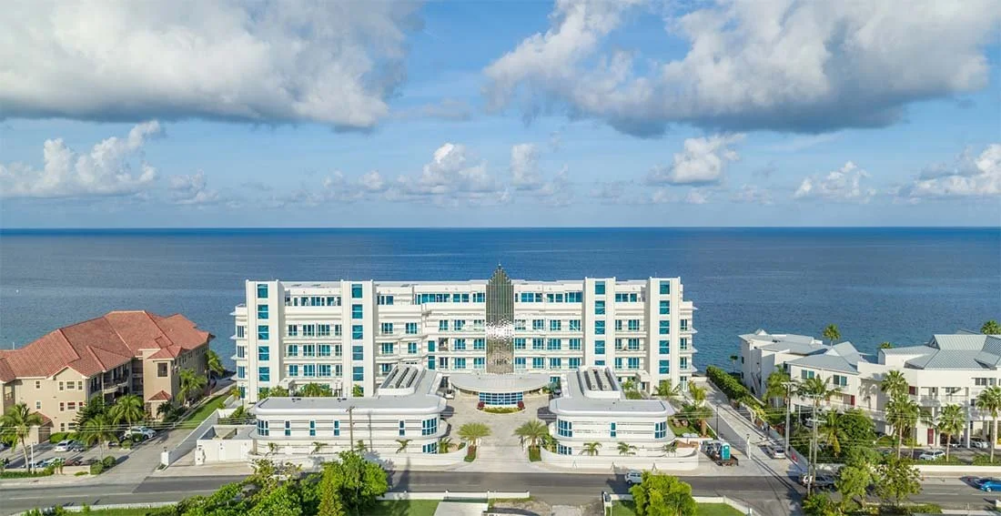 A hotel building near the ocean with a large tower in the center, surrounded by palm trees and smaller buildings, under a partly cloudy sky.