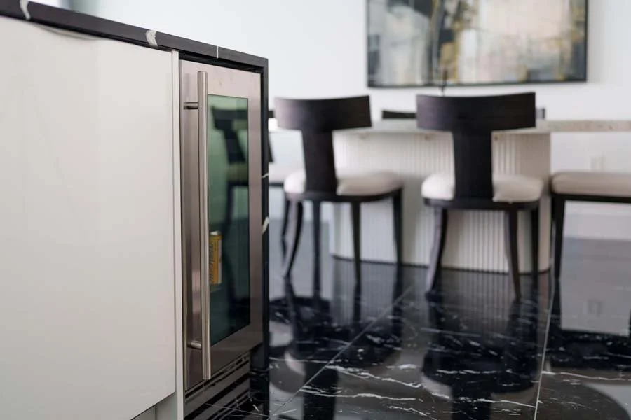 Part of a kitchen with a white cabinet and a small wine or beverage fridge, with black marble floor and dining area with black chairs and a white table in the background.