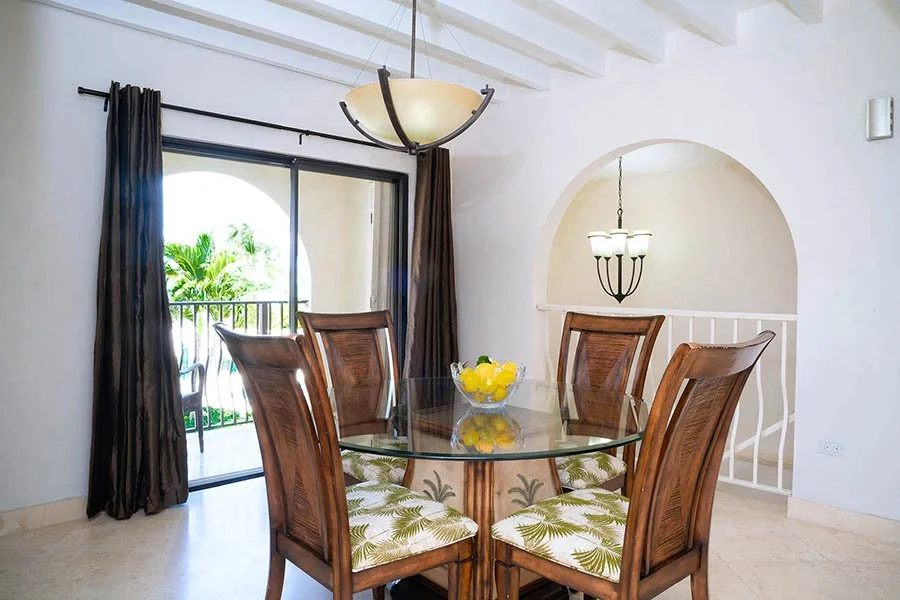 Dining room with a round glass table, four wooden chairs with leaf-patterned cushions, a bowl of lemons, large window with dark curtains, and two hanging chandeliers.