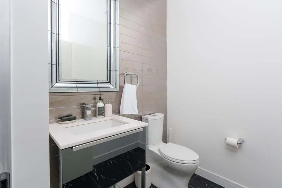 Modern bathroom with a white toilet, a sink with soap and lotion bottles, a towel hanging on a hook, a mirror, and a window, with beige and black tiled flooring.
